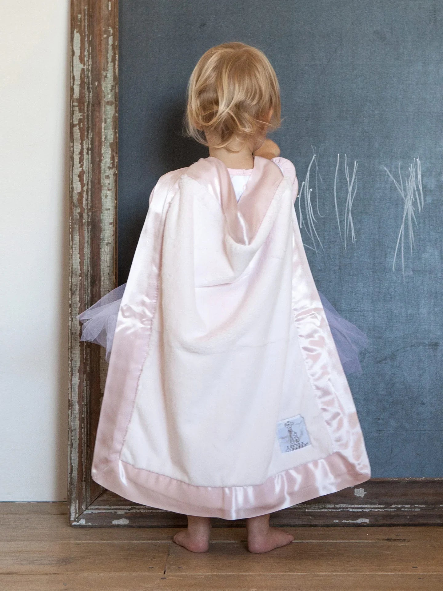 A small child with a pink plush baby blanket wrapped around her shoulders like a cape. The child is making marks on a wood-framed chalkboard siting against a white wall on a wood-paneled floor.